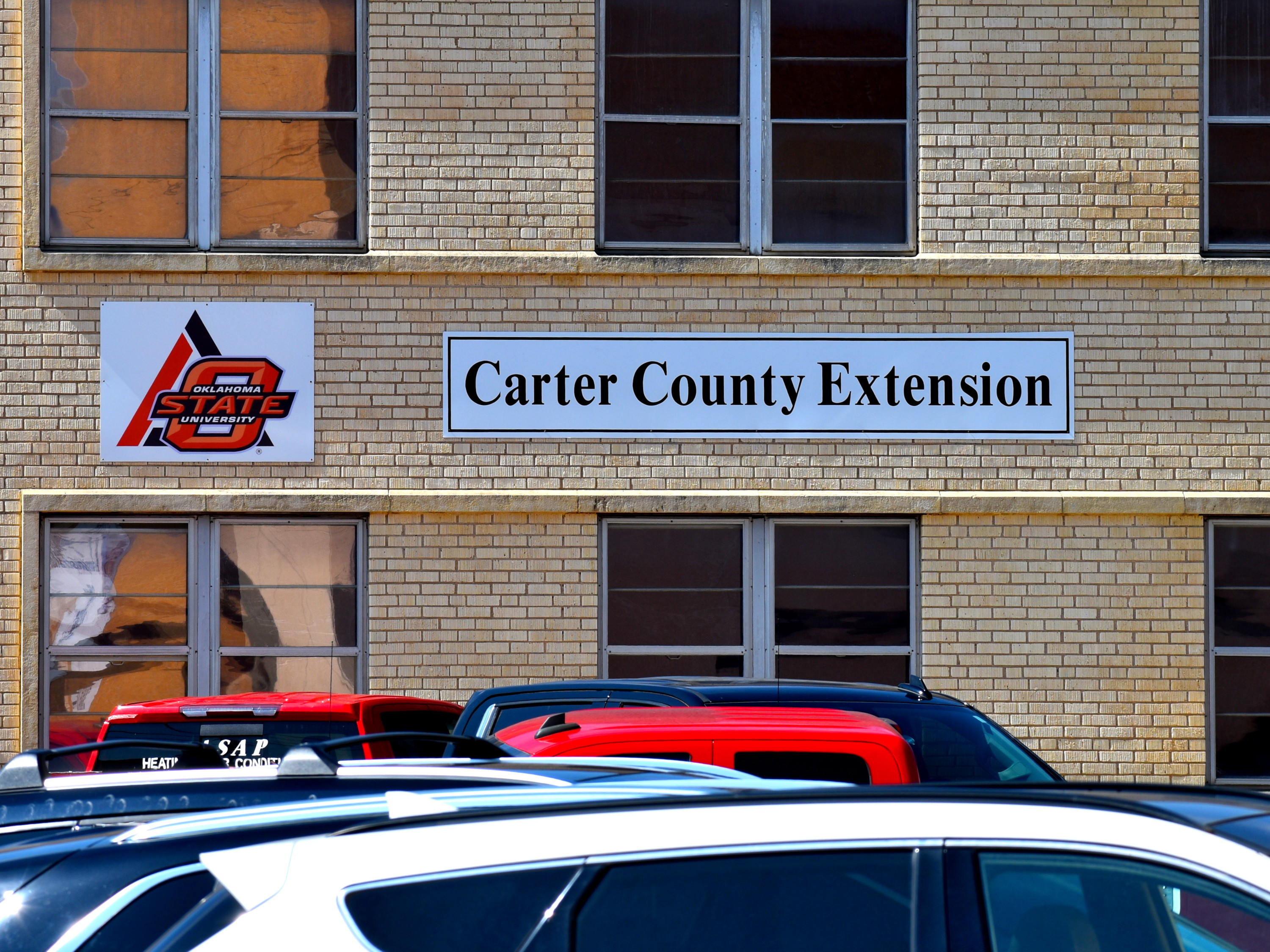 A light brown brick building with the Oklahoma State University logo in orange to the left and a white sign with black letters for the Carter County Extension office. 
